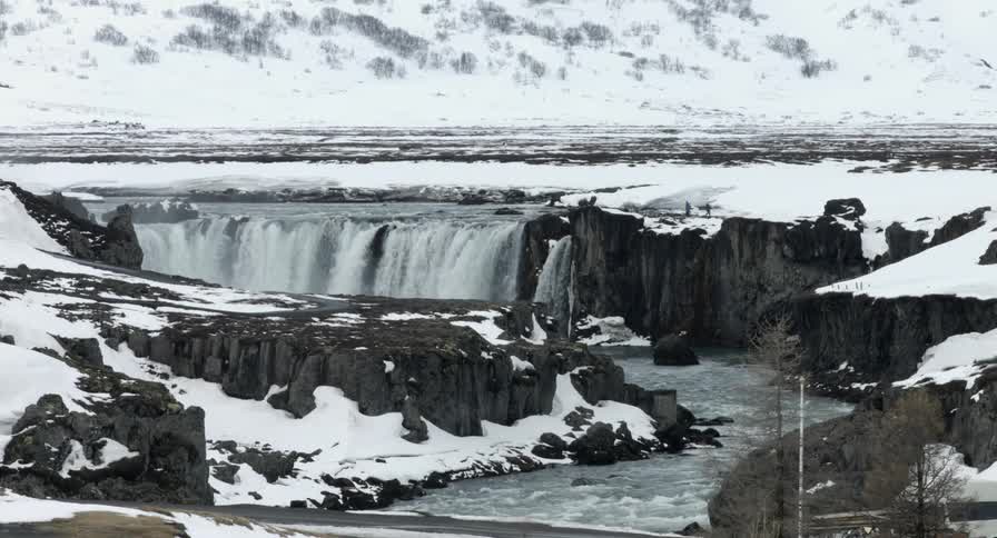 Two people in the distance walk toward a waterfall flowing in a snowy landscape