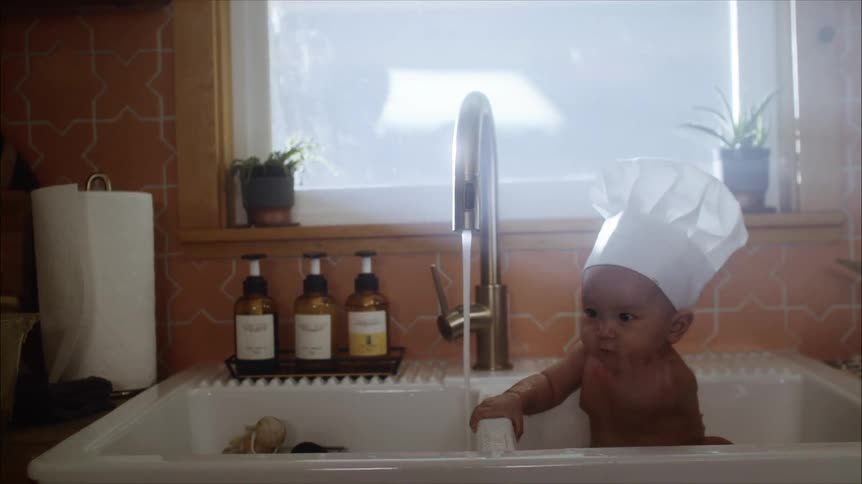 Baby wearing a white chef hat takes a bath on the kitchen sink at home