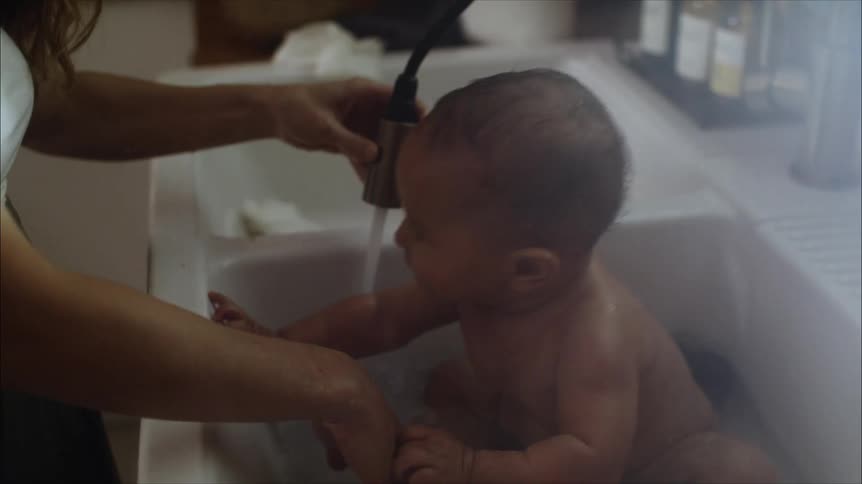 A mother bathing her infant in a white porcelain kitchen sink