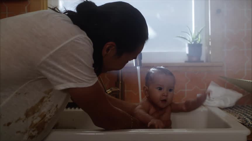A young baby splashes playfully as it is bathed in the sink by an adult