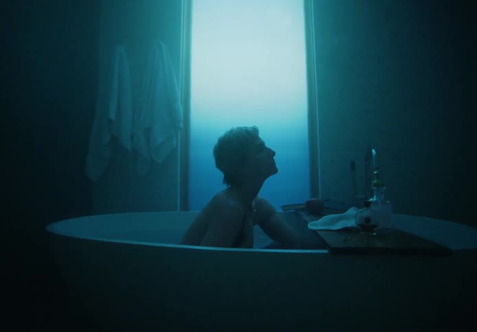 A man is taking a bath in a dark bathroom with light blooming from the covered window in the daytime