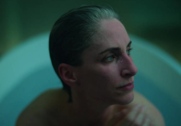 A woman in a bathtub runs her hands over her hair and then rubs her eyes and looks off in the distance