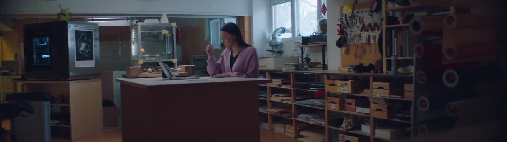 Woman working inside a worksop in front of a laptop near 3d printers and shelves filled with materials