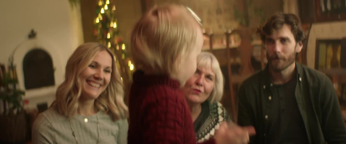 Grandmother and parents watch child jingle bell with christmas tree in the background