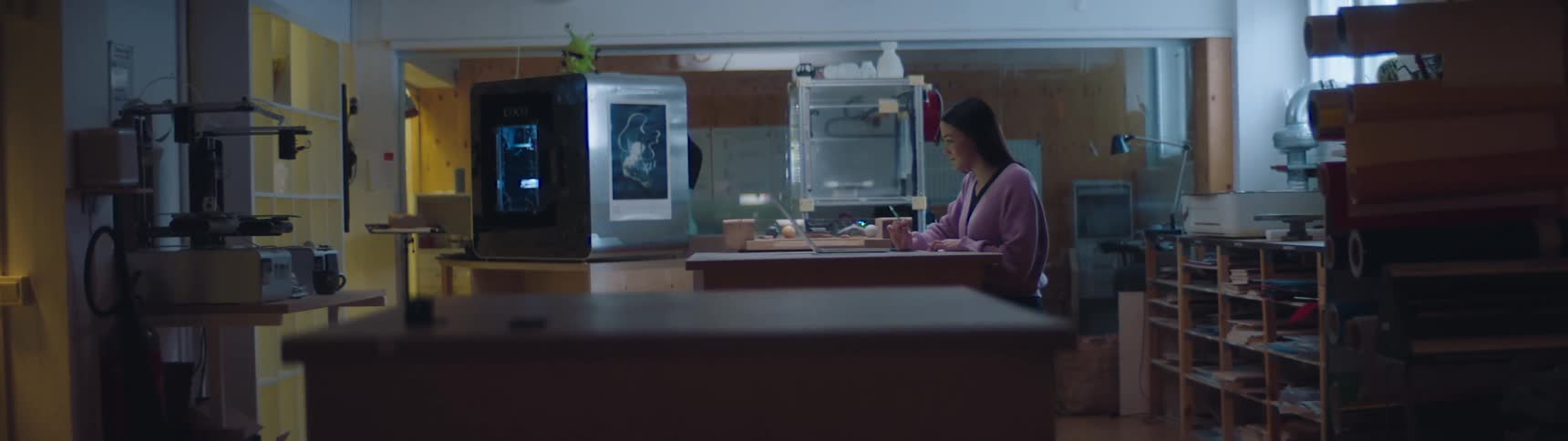 Woman working inside a worksop in front of a laptop near 3d printers and shelves filled with materials