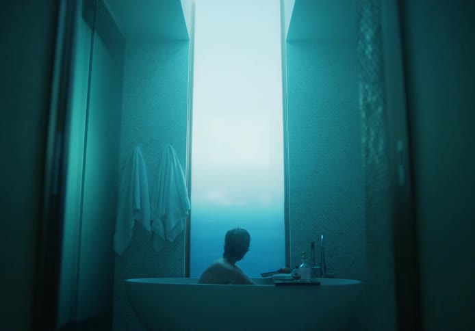 Man sitting in bathtub in bathroom lit by blue tinted light takes a bite of an apple