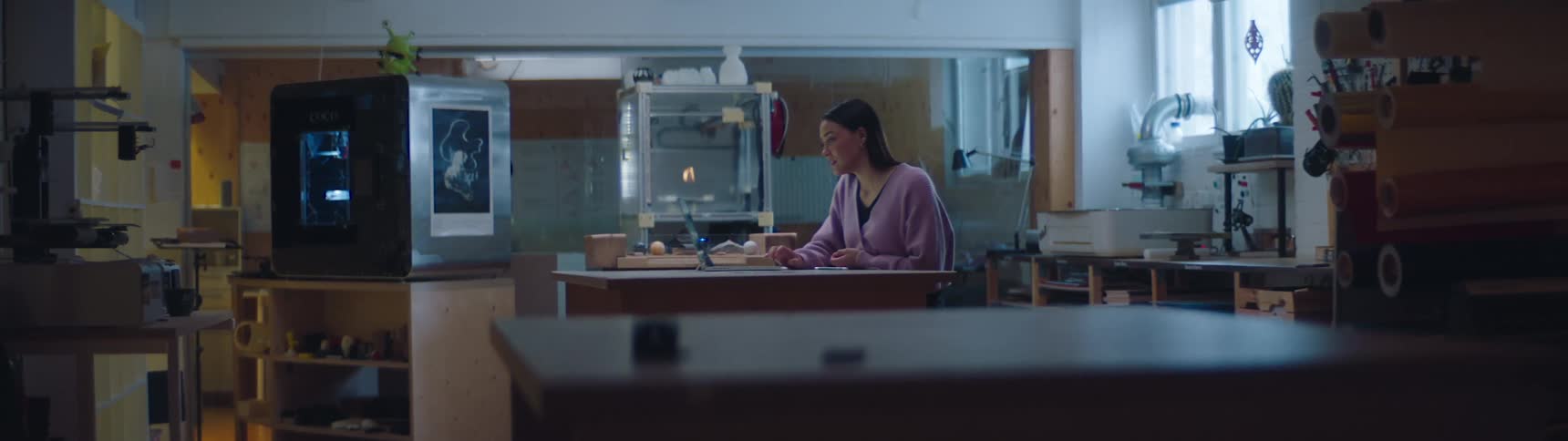 Woman working inside a worksop in front of a laptop near 3d printers and shelves filled with materials