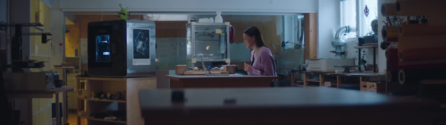 Woman working inside a workshop answers a video call on a laptop near 3d printers and shelves filled with materials