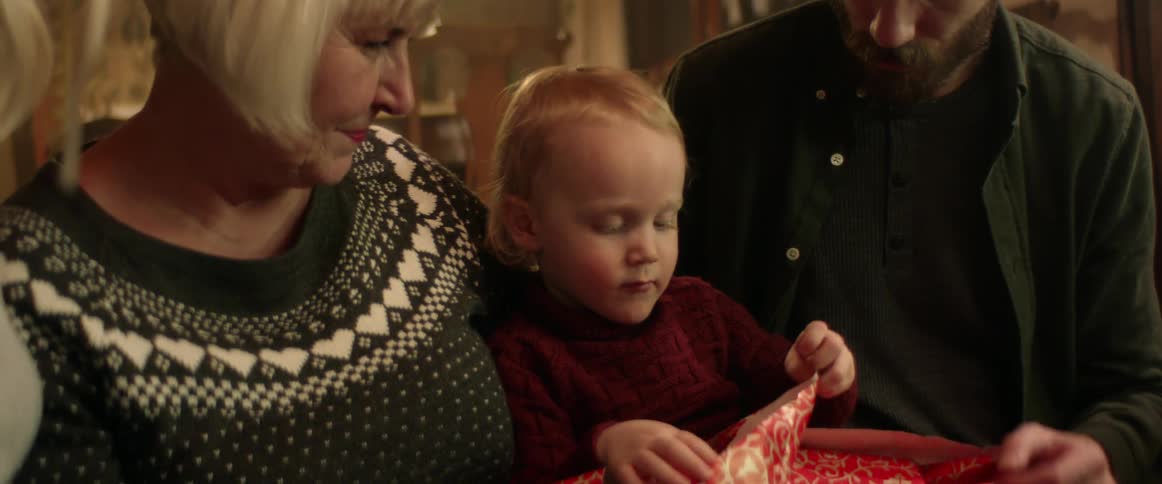 A family is sitting indoors while a child happily unwraps her gift