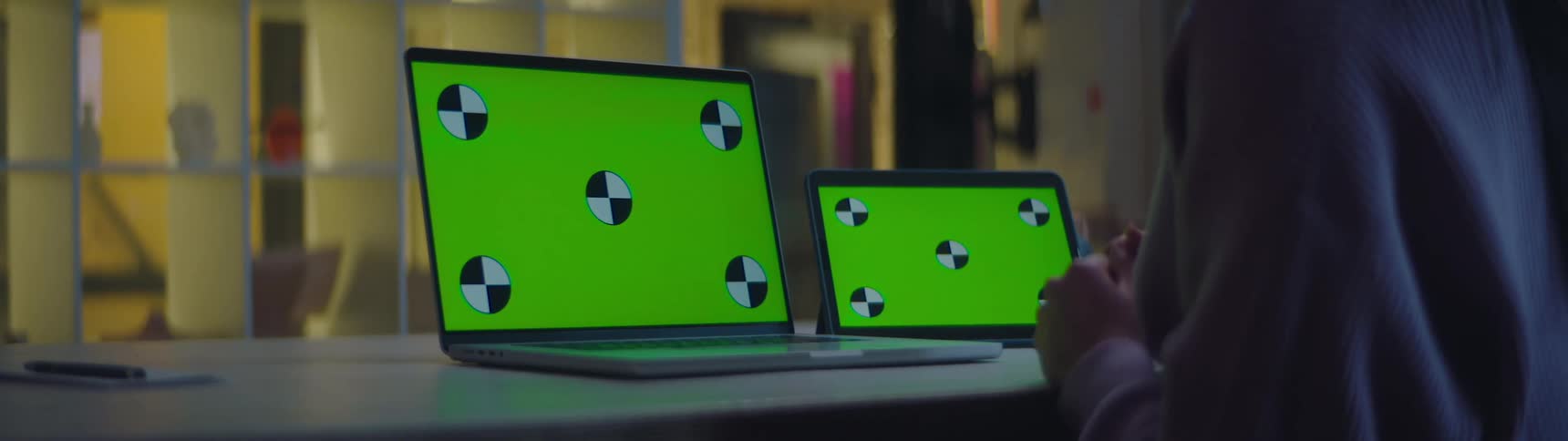 A man is sitting at a desk in an office building with a laptop and tablet showing replaceable green screens