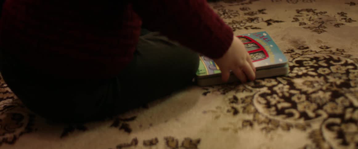 A toddler sits on a rug at home and opens up a book