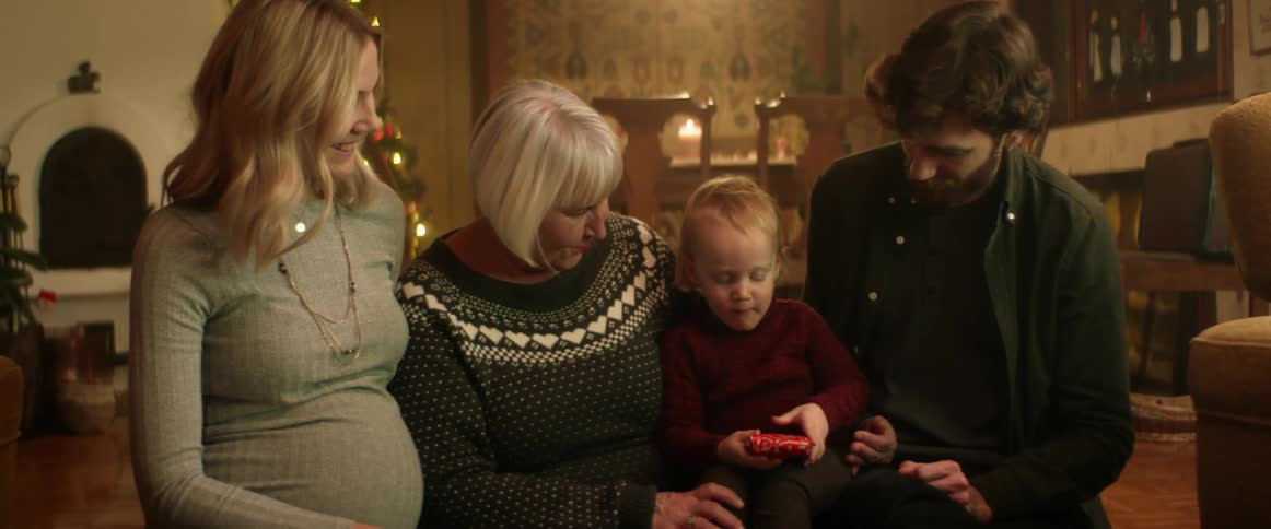 Mom and grandma laugh as they watch dad help his son unwrap a present