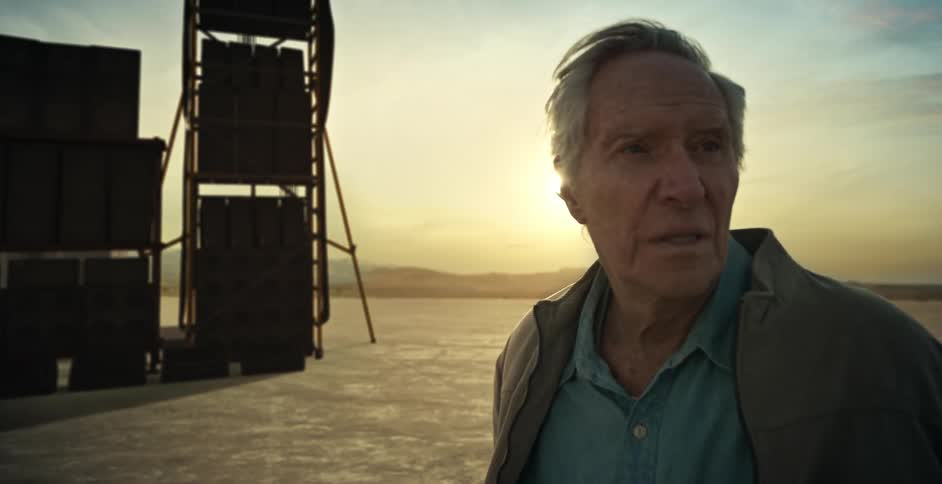 An elderly man is standing outdoors in an open landscape