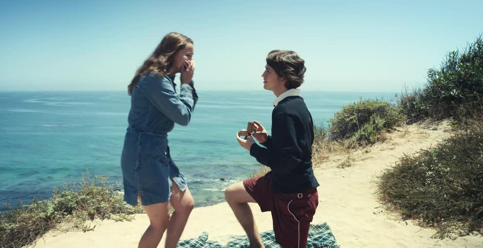 A young man kneels down on the shore and opens a ring box as he proposes to a woman who covers her mouth and hides a scream and hugs him as he stands up to hug her
