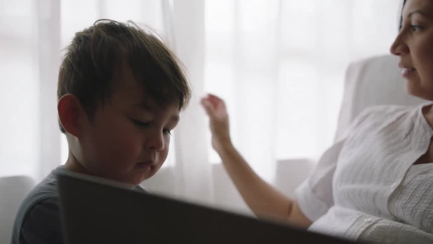 Pregnant mother with white dress sits in armchair and plays with son next to windows with curtains