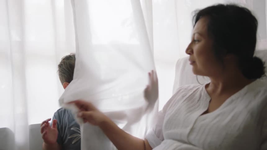 Mother sitting on a couch uses the white curtains to play with her son at home
