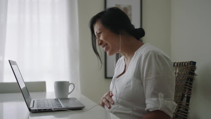 A pregnant woman is sitting at a table in front of a laptop computer waving to someone in a virtual conversation