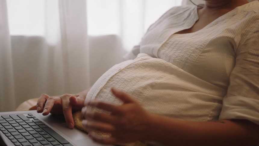 A pregnant woman has a laptop computer in her lap and types for a while and puts her hand on her belly