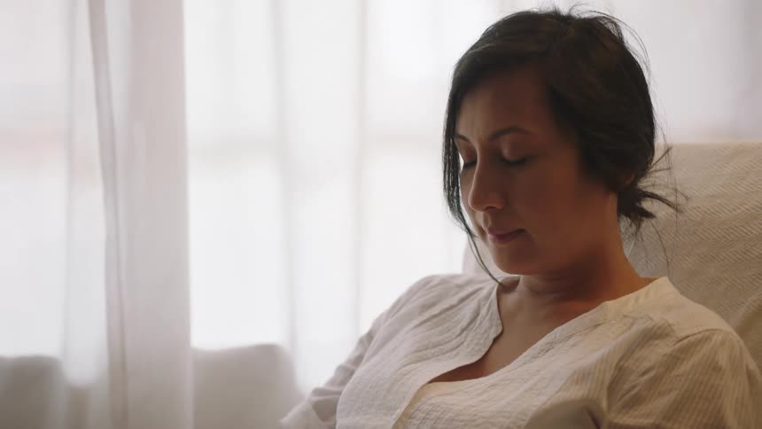 Pregnant woman with a laptop on her lap sitting on a chair at home next to a window