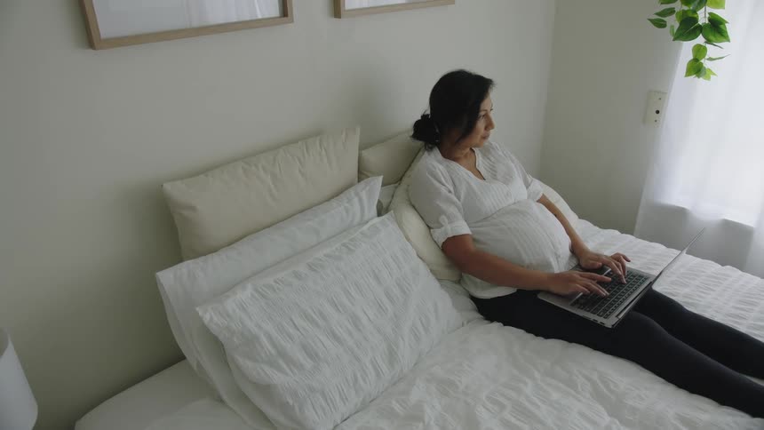 A pregnant woman works on her laptop while lying in bed