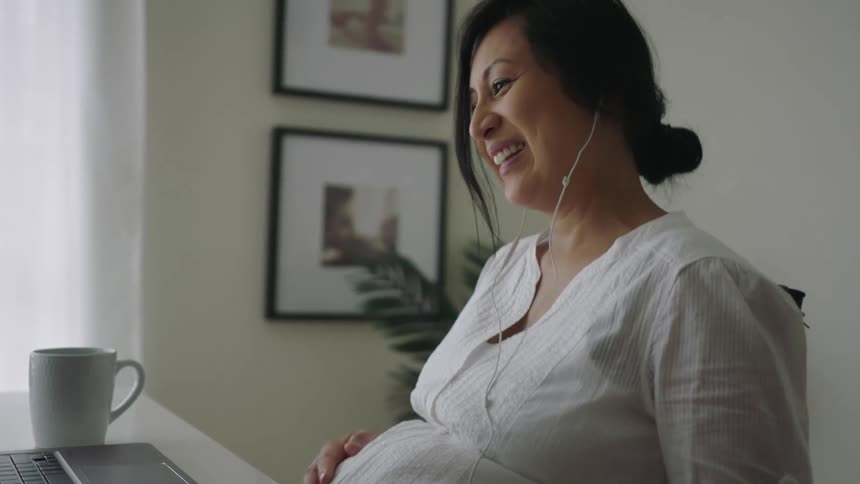 Pregnant woman with earphones and white shirt sits at table making video call on laptop rubbing belly