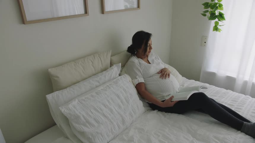 Pregnant woman reading a book while resting in bed in the bedroom at home