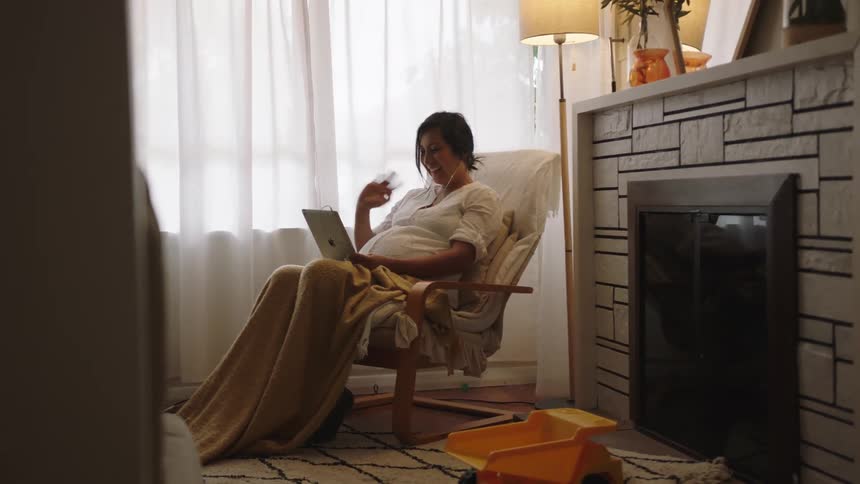A pregnant woman sitting in a chair near a window waving at her laptop when her son walks into the room