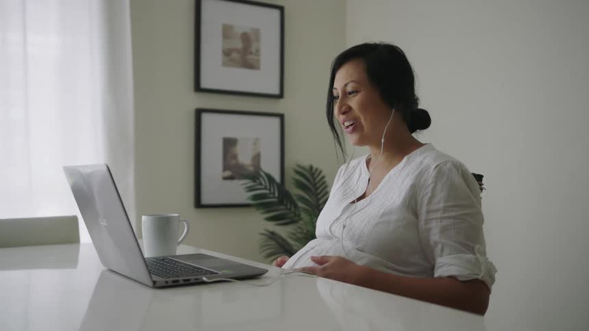 A pregnant woman waves during a video call on her laptop