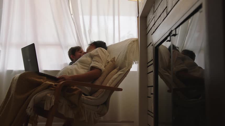 A boy kisses his mother on the lips while the woman works on her laptop at home
