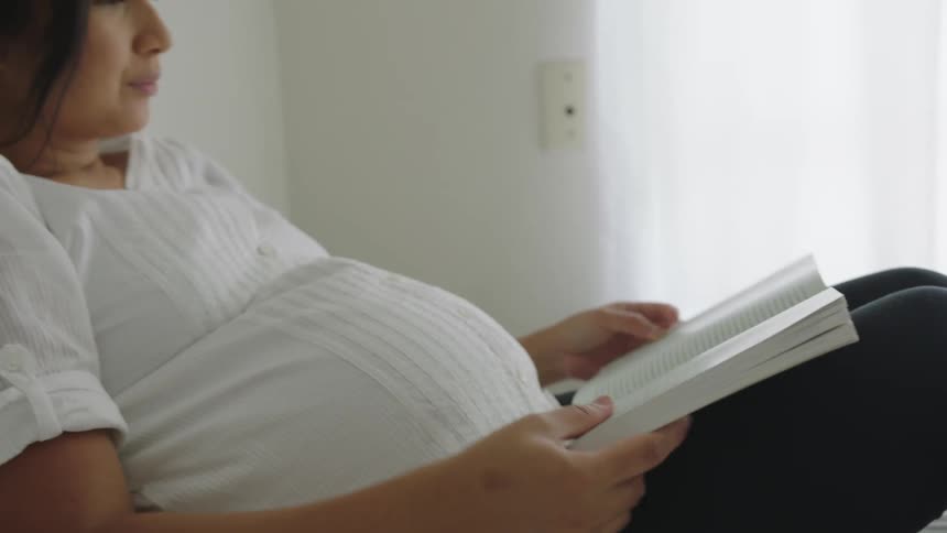A pregnant woman touches and caresses her baby belly while reading a book in bed
