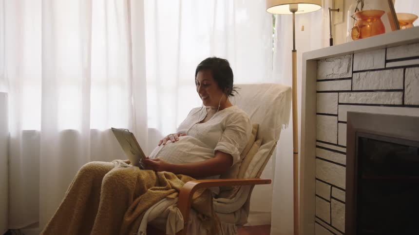 A pregnant woman uses a tablet for a video call while sitting at home