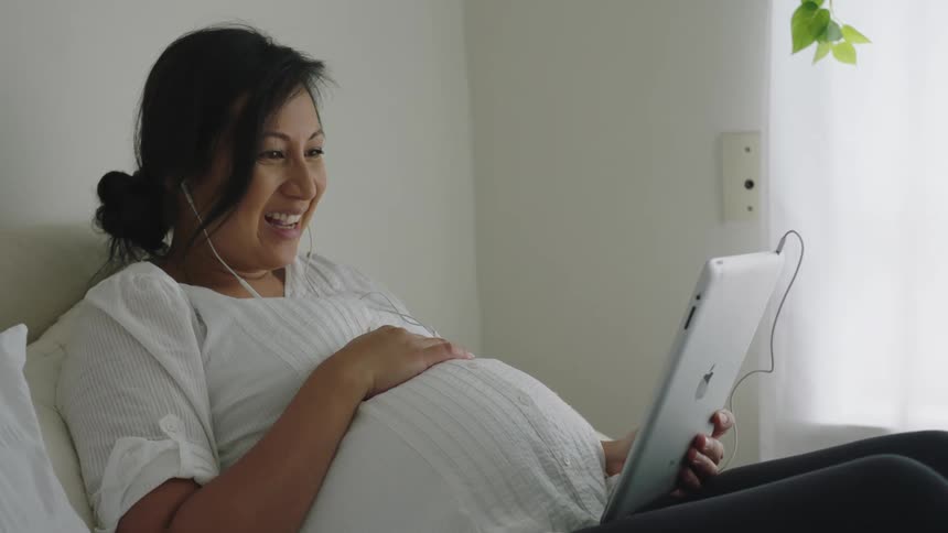 Pregnant woman talking on a video call on a tablet while resting in bed in the bedroom at home