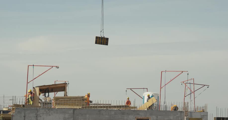 Construction workers stand on a building site as a construction crane lowers material