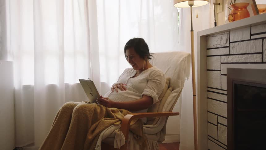 A pregnant woman sits at home and takes her hand off her belly as she waves at her tablet while video chatting
