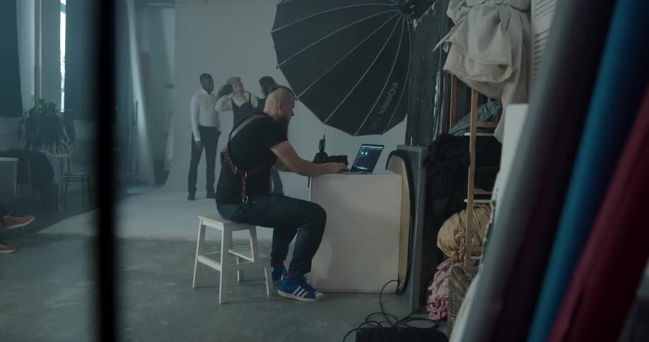 Photographer sits with laptop as dressers adjusts clothing on fashion models standing near backdrop
