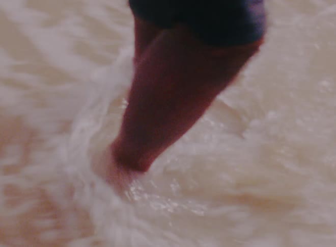 A boy is standing on the sand of a beach while a wave rushes in and covers his bare feet before receding during the daytime