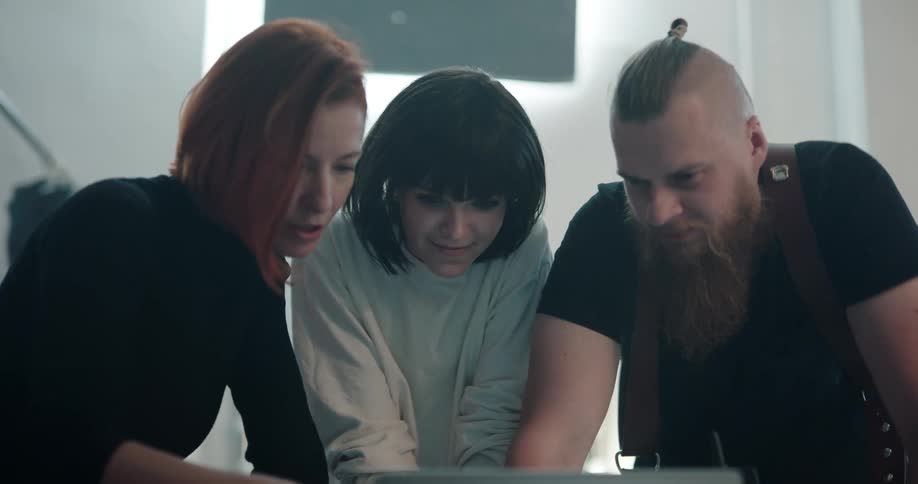 A man with a partially shaved head talks and laughs with two women as they all look at a laptop screen
