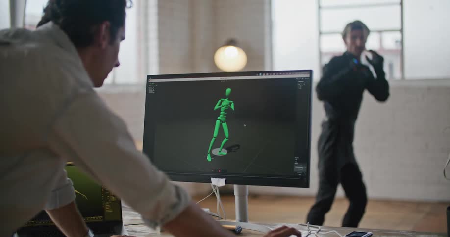 A man wearing virtual reality devices is doing martial arts movements and another man is watching on a computer monitor