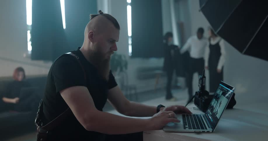 A photographer edits photos on a laptop as a photoshoot takes place behind him