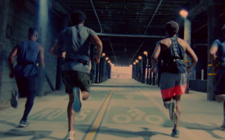 Four men run in tandem on an indoor track under bleachers