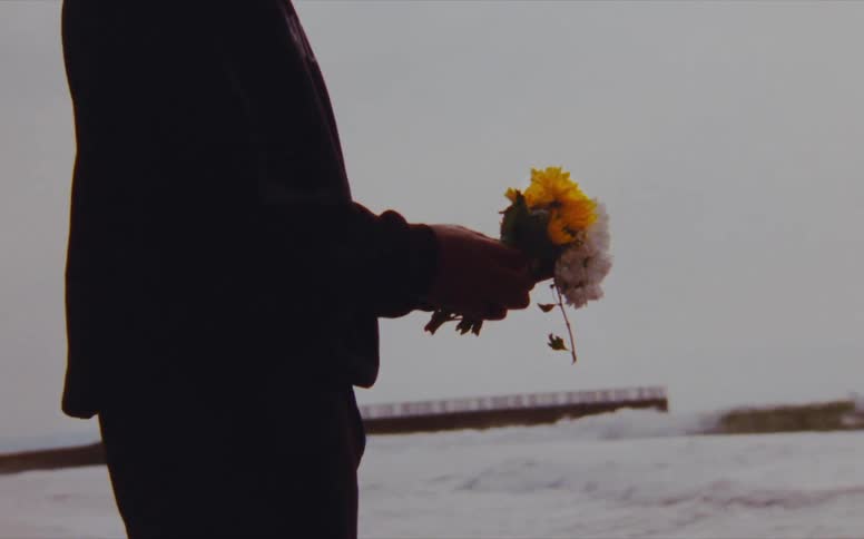 A man is standing on a beach holding a small bouquet of flowers while foaming waves crash near him on an overcast day