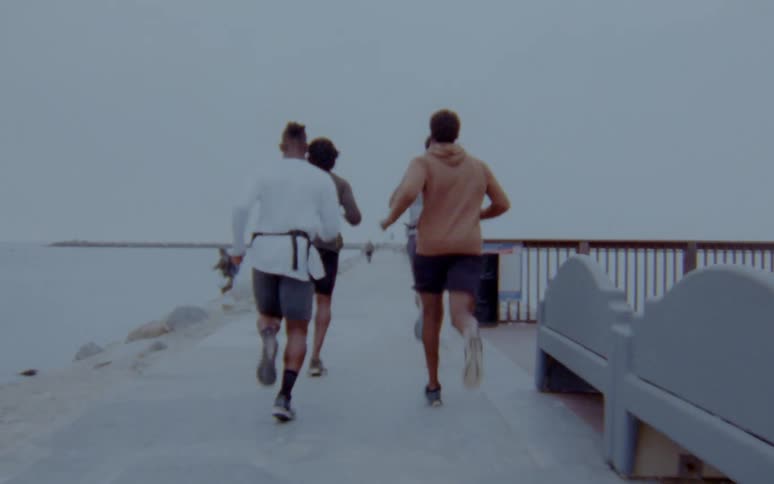 Four men run along the shore past a person fishing on the bank