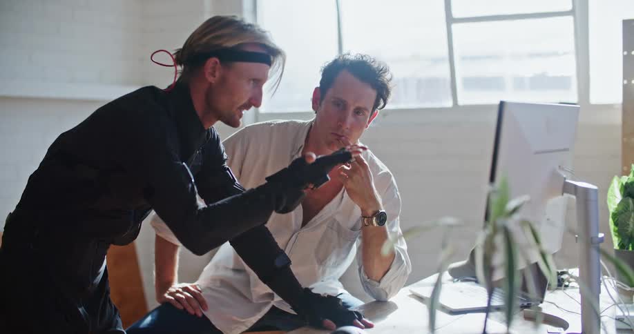 A man has interactive computer virtual reality devices on his hands and is moving his hand as he speaks to a man sitting behind a computer