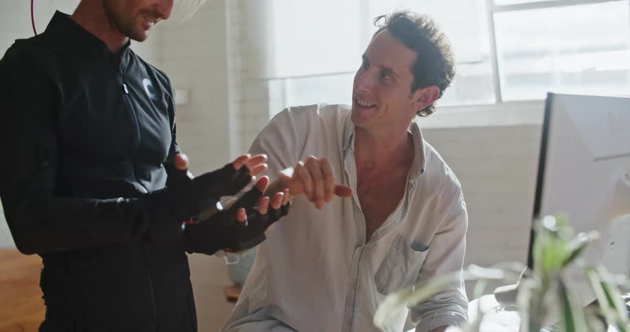 A man has virtual reality devices on his hands and is moving his hands as he speaks to a man sitting behind a computer