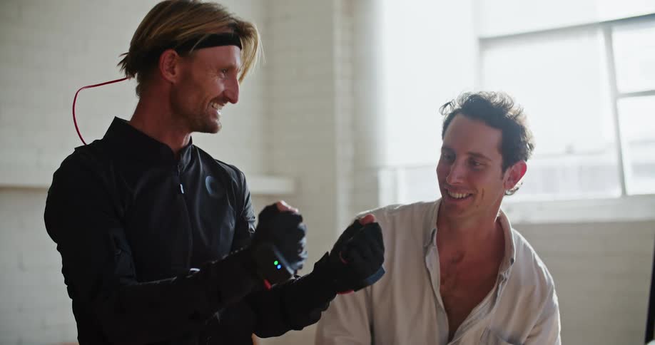 Man wearing virtual reality gaming gloves talks about them to another man as the other man points to a computer monitor
