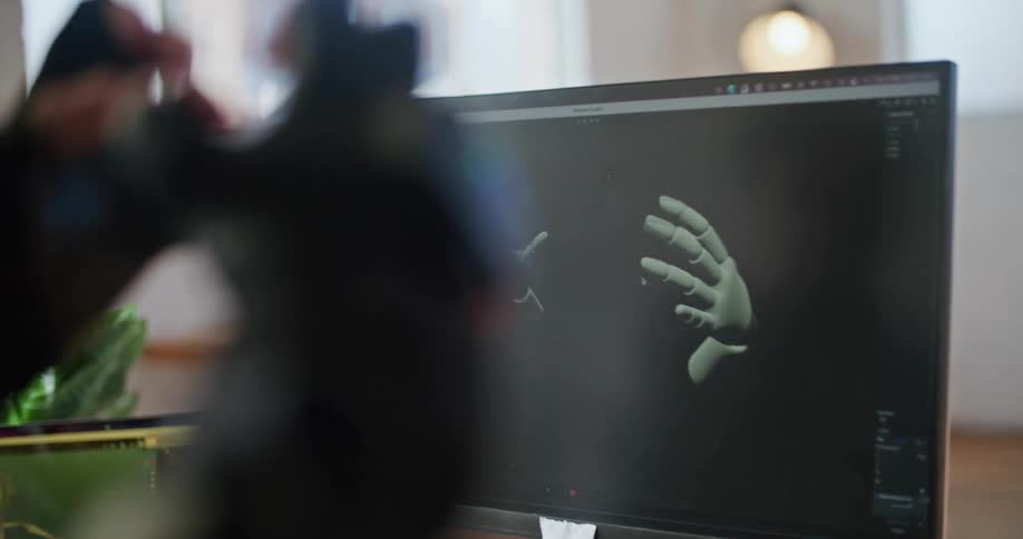 A man in an office with a device strapped to his hands watches a computer in front of him mimic his hand motions as another man watches