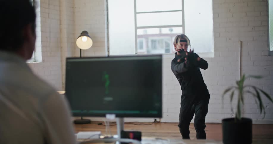Man wearing a black motion capture suit moving into fighting poses inside a white room in front of a computer