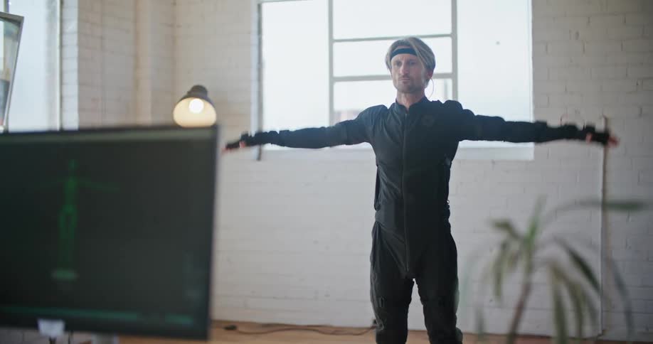 A man wearing virtual reality devices stands with his arms to his sides and then lifts them as someone looks at a computer monitor