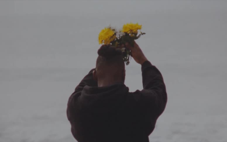 Man holds yellow flowers above his head before he lowers them as he faces the water