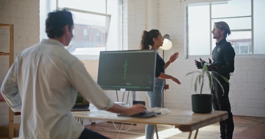 A man wearing virtual reality devices is speaking with a woman as a man is sitting behind a desk with computers